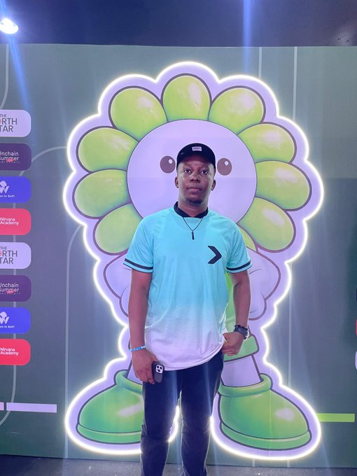 A man wearing a light blue and white t-shirt, black cap, and jeans stands in front of a large illuminated display of the Takashi Murakami flower design.