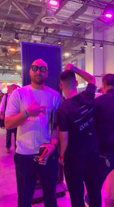 A bald man wearing distinctive diamond-shaped sunglasses and a white t-shirt poses with a hand gesture at an indoor event with purple lighting, while another person in a black 'ZUKTY' shirt stands nearby.