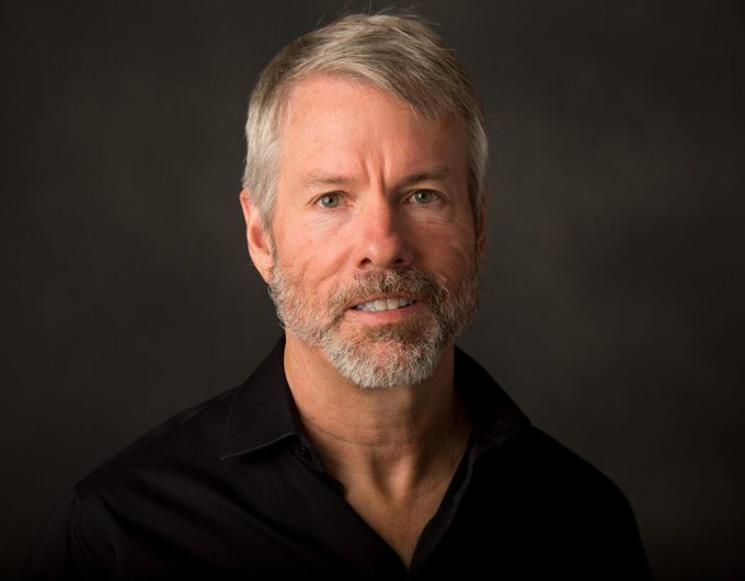 A professional headshot portrait of a gray-haired, bearded man wearing a black shirt against a dark gray background.