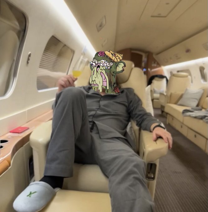 A person in gray loungewear relaxes in a luxury private jet cabin, holding a glass of yellow liquid, with their face replaced by a cartoon zombie monster sticker.