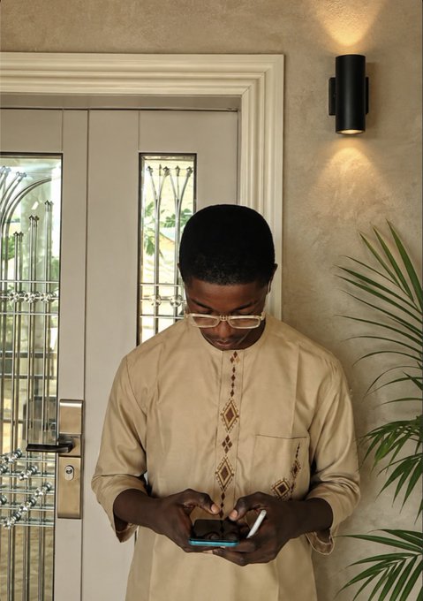 A young man wearing glasses and a beige embroidered tunic stands in a doorway, looking down intently at his smartphone.