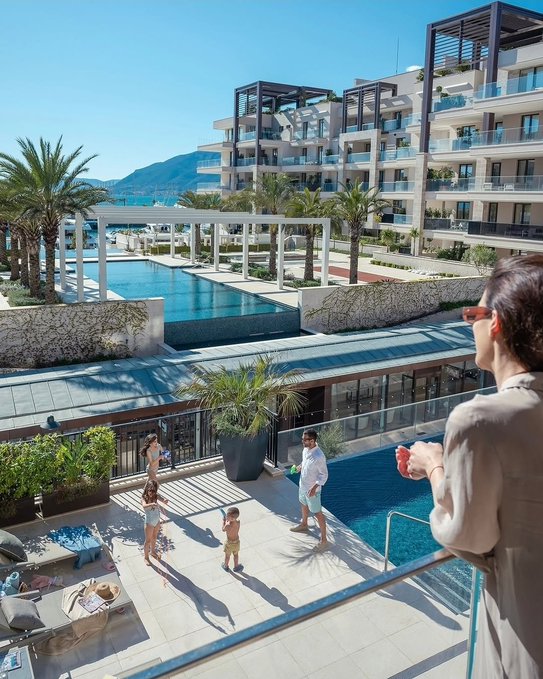 The image shows a woman standing outside, looking at a pool area with several people playing around in the water. There are two children nearby who seem to be enjoying themselves as well. The scene appears to be set in an apartment complex or hotel, where guests can relax and have fun by the poolside.