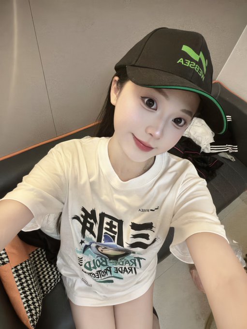 A selfie of a young woman wearing a white graphic t-shirt with Japanese and English text and a black cap with green 'NEBULA' lettering.