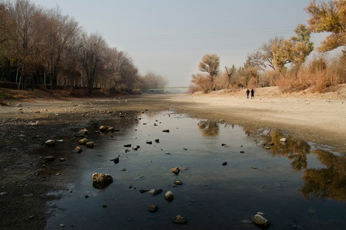 The image shows a river with two people walking along its edge, enjoying the serene surroundings. The river appears to be shallow, as there are rocks scattered around it. In the background, trees can be seen lining the riverbank, adding to the picturesque scene. The photo is posted on social media and has been tagged by @LeslieASpurlock, who wrote a message expressing gratitude for the image being in their collection.