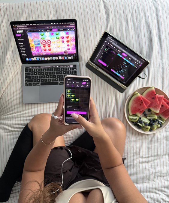 A first-person view of someone on a bed holding a smartphone with a music app open, surrounded by a laptop, tablet, and a plate of watermelon slices, with headphones around their neck.