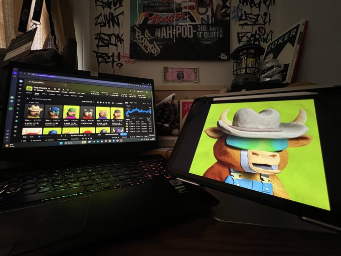 A dimly lit desk setup showing a tablet displaying an NFT marketplace and a laptop screen featuring a cartoon cow wearing a cowboy hat.