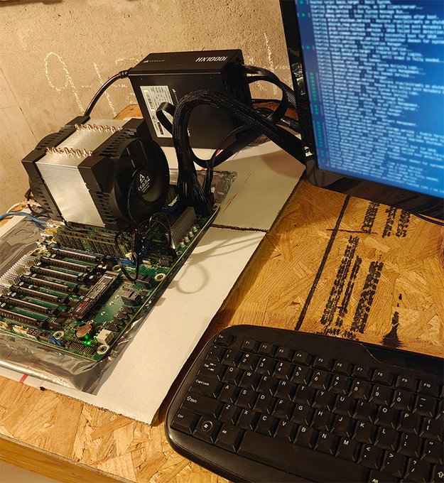 A makeshift open-air computer setup featuring a motherboard with a large CPU cooler and a "NEXTODE" branded power supply unit sits on a wooden surface next to a monitor displaying code and a black keyboard. The DIY build, lacking a traditional case, illustrates the hands-on assembly process of the user's experimental low-power Theta Network EdgeCloud hobby inference server mentioned in the post.