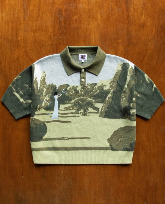 A folded short-sleeve knit polo shirt featuring a green-toned tapestry-style landscape print with a bride, laid flat on a wooden surface.