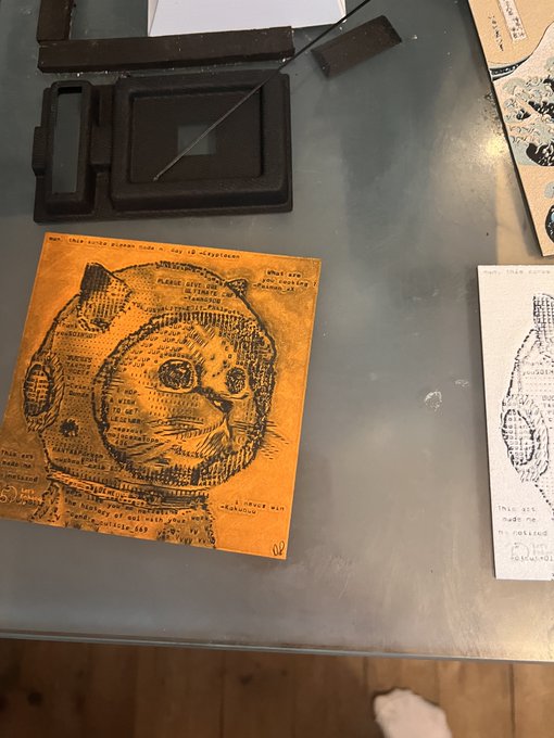 A work-in-progress art piece on yellow paper depicts a creature resembling a bear or cat in a space helmet, created using text from a printed page. Black printing blocks sit at the top of the workspace.