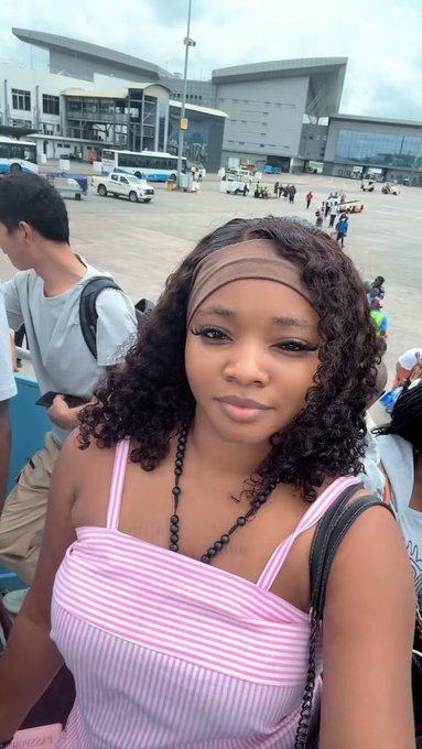 Selfie of a young woman with curly brown hair wearing a pink and white striped top and a beaded necklace, taken outdoors near what appears to be an airport or transit terminal.