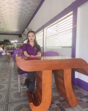 A woman in a purple uniform stands behind a wooden table inside a brightly lit room with purple trim and patterned tile flooring.