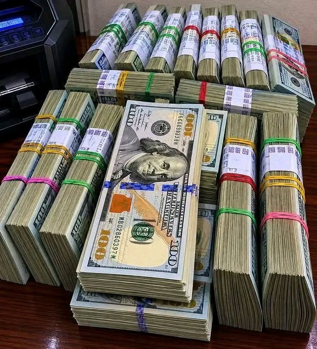 Stacks of $100 US dollar bills bound with colorful currency straps are arranged on a table next to a money-counting machine.