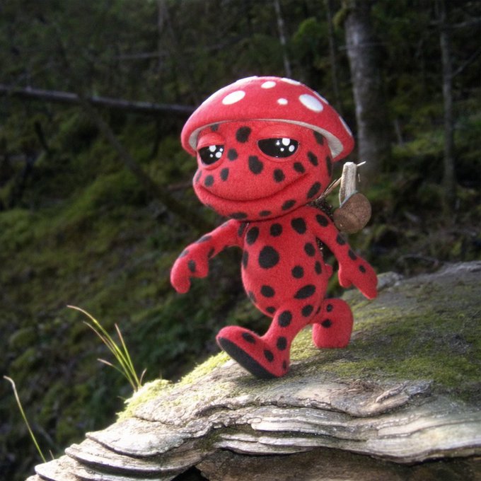 A plush toy of a red, spotted mushroom creature with large anime-style eyes, carrying a small wooden backpack as it walks across a moss-covered log in a forest.