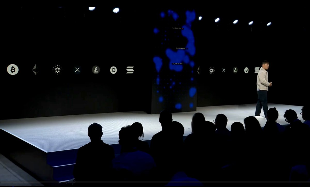 The image shows a man standing at a podium with a large screen behind him, likely displaying information or visuals related to cryptocurrency. The audience consists of people sitting in chairs, attentively watching the speaker. The background features a stage setup with a blue light on one side and a black wall adorned with various logos and icons. One of these logos is for @coinbase, which has enabled users to access loans against their Staked Solana holdings without having to unstake their holdings first.