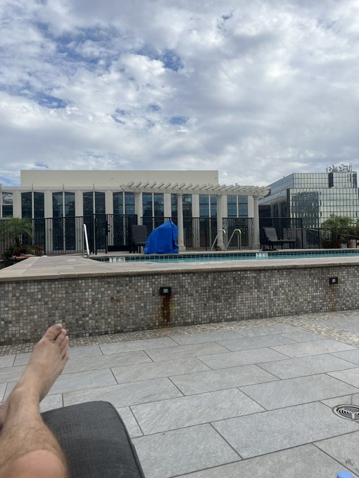 A photo taken from a lounge chair by a rooftop pool, showing a person's legs and feet, with a modern building featuring tall glass windows and a small blue umbrella in the background under a partly cloudy sky.