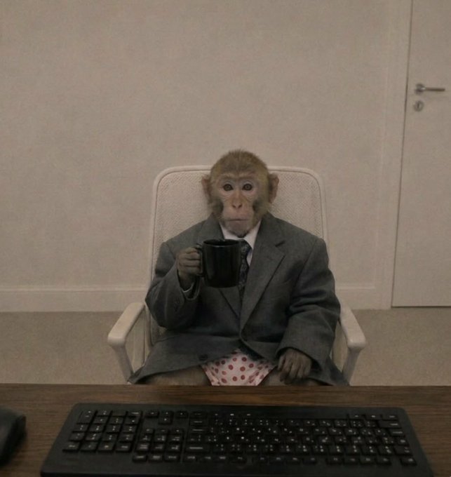 A monkey wearing a gray business suit and polka-dot boxers sits in a white office chair at a desk, sipping from a black mug in front of a computer keyboard.