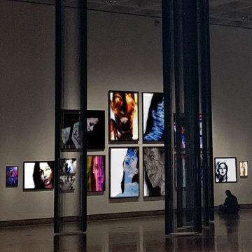 A dimly lit modern art gallery with large black columns, displaying illuminated portrait photographs on the walls while a solitary figure sits on the floor observing.