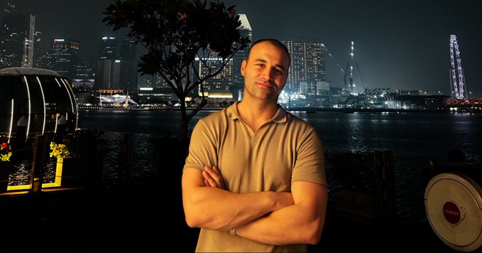A photograph of a muscular man in a tan polo shirt standing with crossed arms against a nighttime city skyline featuring illuminated skyscrapers and waterfront.
