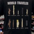 A dark t-shirt displays "WORLD TRAVELERS" in white text at the top, with silhouettes of iconic international landmarks like the Statue of Liberty, Big Ben, the Eiffel Tower, and the Pyramids arranged on a world map, paired with the post asking "send route wen columbia?" as a playful nod to exploring travel destinations.