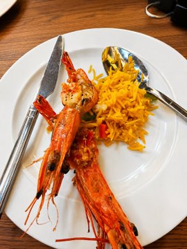 The image features a plate of food, including two cooked prawns with orange shells sitting on top of a white plate. The plate also has a fork and spoon placed next to it. In the background, there are some blurred details that suggest a dining table or countertop setting. The post appears to be about an event where someone purchased tickets for a dinner at a restaurant called "Loshmi shilled so hard that i bought and booked P within a day."