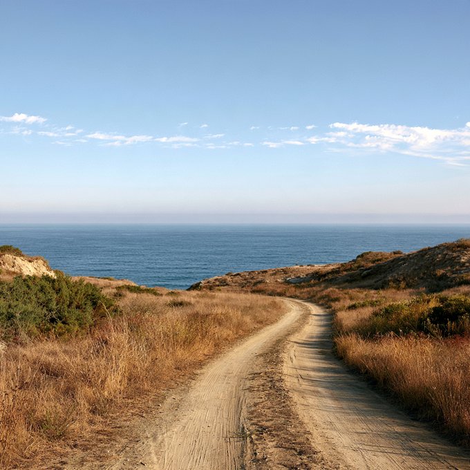 A dirt road winds through dry grassy hills toward the ocean under a blue sky with scattered clouds.