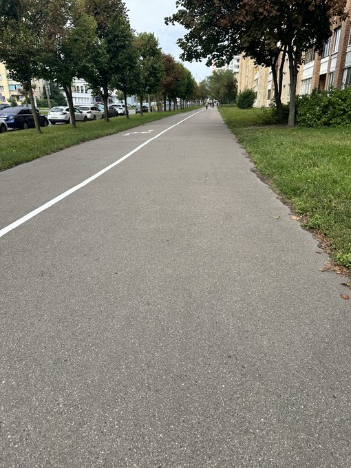 A view down a paved pedestrian path or bike lane lined with trees, parked cars, and buildings, with a few people walking in the distance.