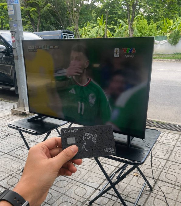A hand holds a KAST Visa card in front of a TV streaming a football match on VTV6 (showing a player in a green Mexico jersey #11), illustrating the post's theme of enjoying football while the KAST card enables everyday cashback rewards.