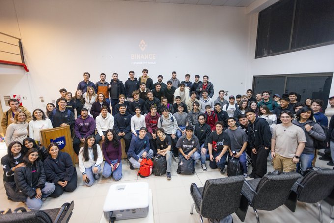 A group of approximately 90 university students pose for a group photo in front of a "BINANCE" branded backdrop, gathered at Universidad Champagnat for the final stop of the Binance University Tour in Mendoza. The students, seated and standing together in a large hall, reflect the enthusiastic turnout described in the post, capturing the close of the crypto education tour.