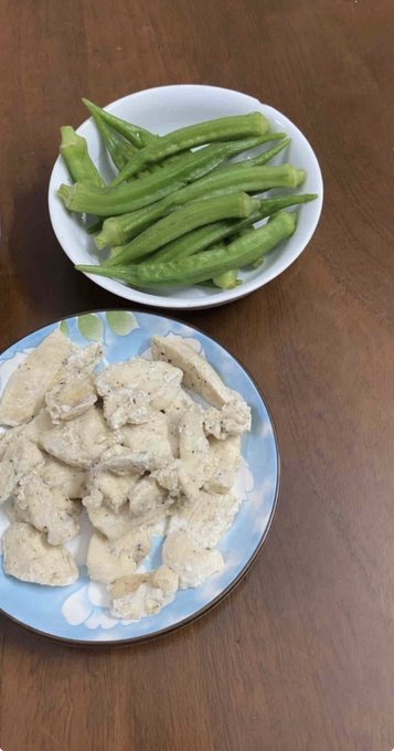 Two plates of food on a wooden table: a bowl of fresh green okra pods on top and a blue plate of steamed white dumplings or fish cakes below, with a wedge of lime.