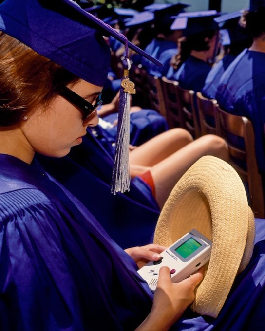 A young woman wearing a purple graduation cap and gown plays a Game Boy while holding a straw hat, seated among other graduates.