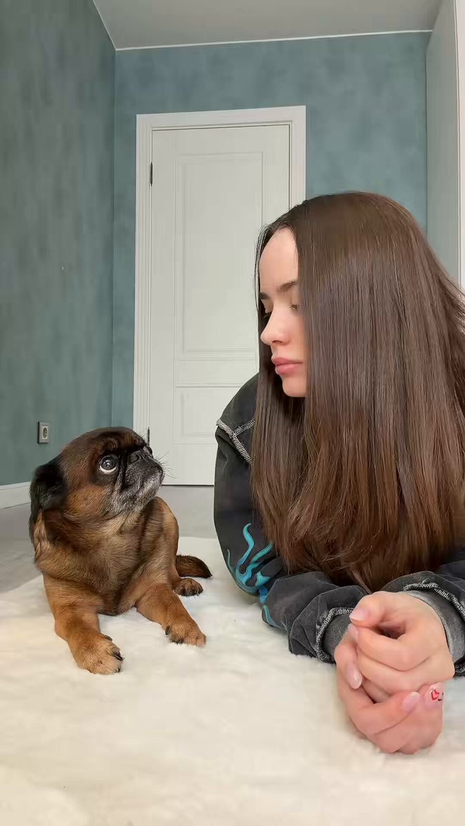 The image shows a woman lying down next to her dog on a white bedspread. The woman appears to be looking at or interacting with the dog while they are both on the floor, possibly posing for a photo.