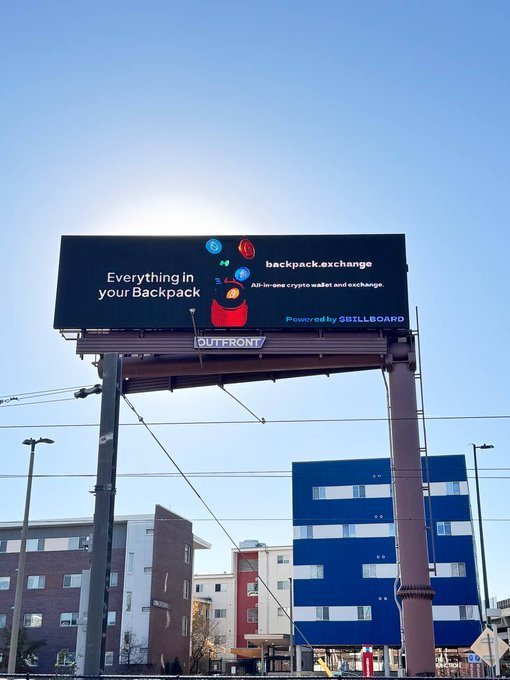 The image shows a billboard with an advertisement for a backpack exchange service, featuring a large sunburst graphic of a backpack flying through the air. The text on the billboard reads "Everything in your @backpack backpack.exchange/signup?referra". This billboard is located near some buildings and appears to be part of a larger urban setting with various architectural styles.