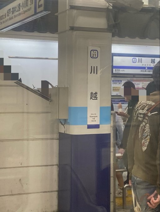 The image shows a subway station with a blue and white pillar, which has a sign that reads "Step Narrow". There are people standing around the pillar, possibly waiting for their train or just passing by. The background of the image features a wall with some writing on it in Japanese characters.