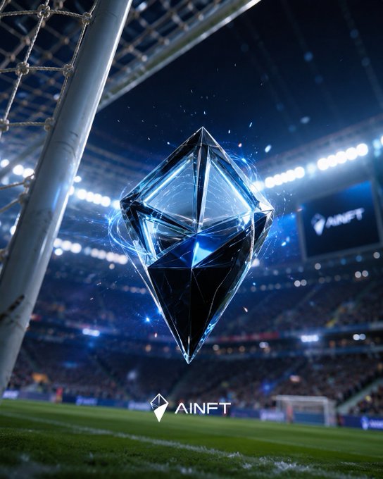 A glowing blue crystalline diamond floats above a soccer stadium field near the goal net, with "AINFT" branding displayed below and on a screen in the background. The image visually represents the post's message about AINFT's fusion of AI and blockchain, with the radiant, transformative crystal symbolizing the dynamic, evolving digital assets and intelligent game characters the platform aims to create within the Web3 ecosystem.