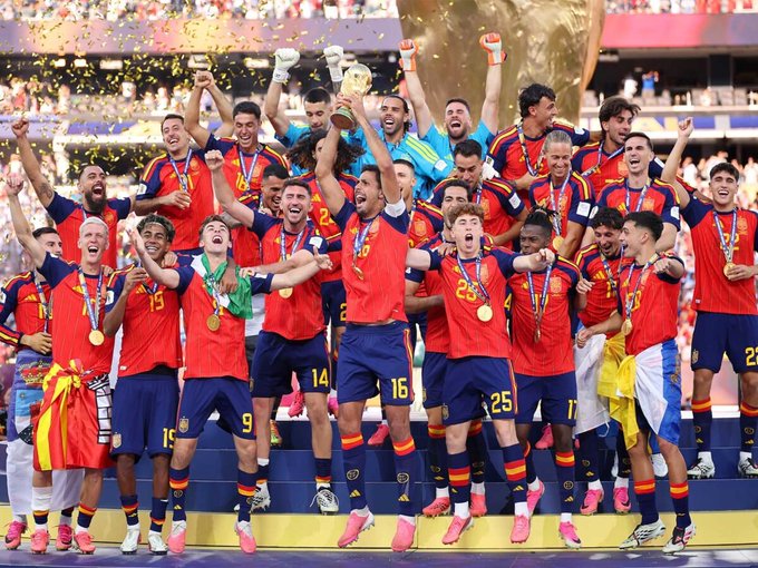 The Spanish national football team celebrates on the podium with a trophy raised overhead and gold medals around their necks, commemorating their victory over Argentina. Confetti rains down as the players, wearing red and blue jerseys with numbers like 9, 14, 16, 22, and 25 visible, cheer in unison following their extra-time triumph.