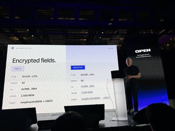 A presenter stands on stage at a tech conference beside a large screen displaying a slide titled "Encrypted fields" that compares public and encrypted blockchain transaction data, with the word "OPEN" visible in the background.
