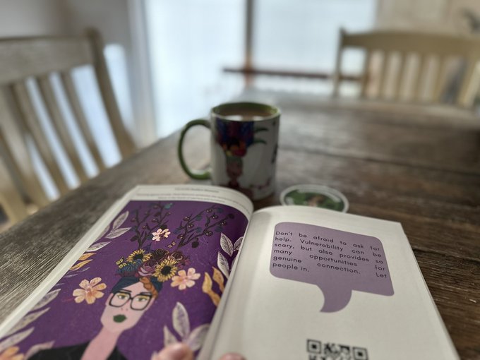 An open illustrated book on a wooden table next to a patterned coffee mug, with a speech bubble reading "don't be afraid to ask for help. Vulnerability can be scary, but also provides so many opportunities for genuine connection. Let people in."