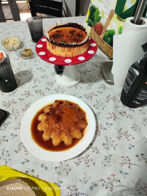 A homemade birthday flan dessert is served on a white plate alongside another whole flan on a polka-dot cake stand, likely prepared for a celebration as suggested by the #cumpleaños (Spanish for "birthday") hashtag. The 1 KG bag of sugar visible on the table hints at the baking process behind the sweet treat.