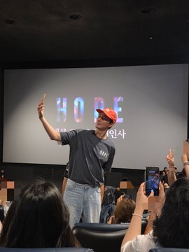 A man wearing a red "HOOD" cap and dark shirt gestures with his left hand while addressing a crowd at what appears to be a movie event in Busan Centum, with the post suggesting this is actor Jo In-sung at the screening.