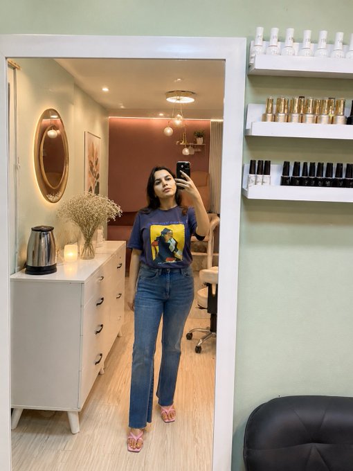 Mirror selfie of a woman wearing a blue graphic t-shirt, high-waisted blue jeans, and pink sandals, standing in what appears to be a salon with shelves of nail polish bottles on the right.
