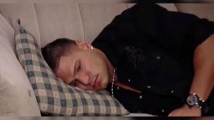 A man with a goatee wearing a black shirt and silver chain is sleeping on a pillow with a checkered pillowcase.