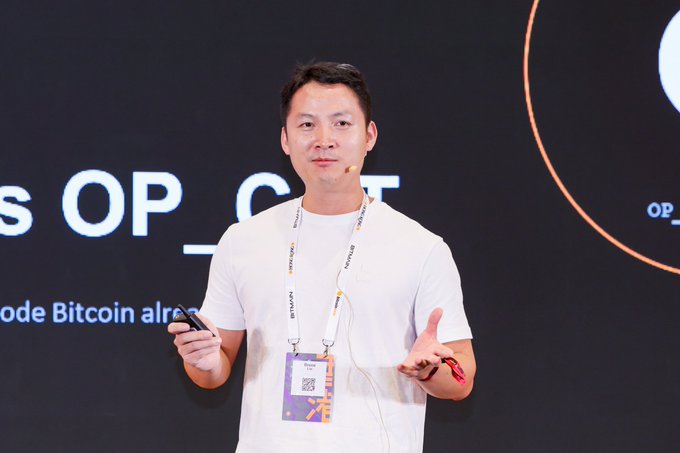 An Asian man in a white t-shirt wearing a 'VIP Club' lanyard speaks on stage with a microphone, holding a remote, with partial text 'OP_CAT' and 'node Bitcoin already' visible on the screen behind him.