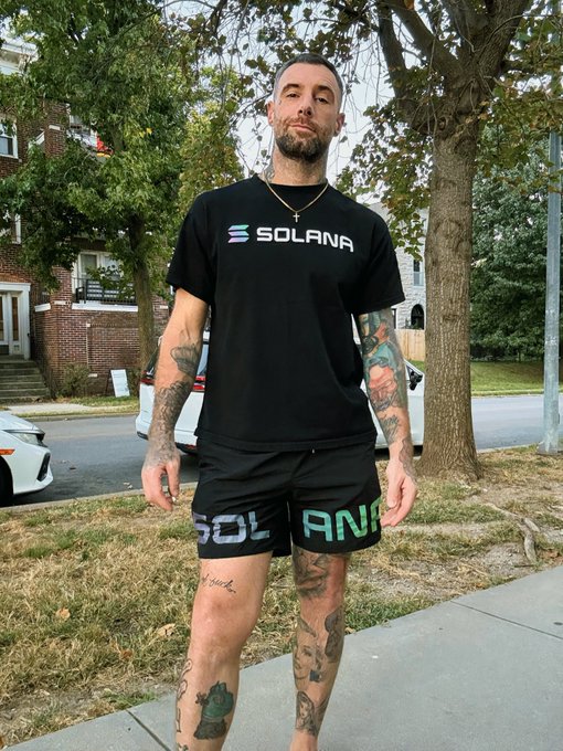 A tattooed man standing outdoors wearing a black 'SOLANA' t-shirt and matching 'SOLANA' shorts with iridescent lettering.