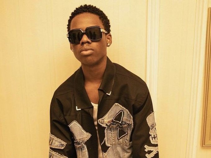 A portrait of a young Black man with short hair, wearing dark sunglasses, diamond earrings, and a black denim jacket with denim patches over a white shirt.