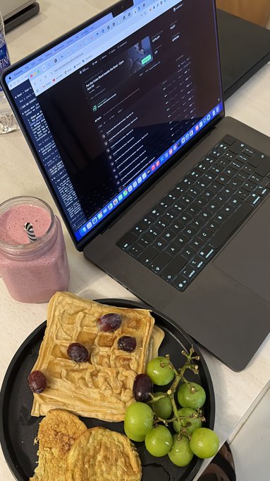 A laptop displaying trading charts and data sits beside a breakfast plate of golden waffles topped with grapes and a pink berry smoothie, pairing the post's discussion of Axis Robotics' $12M raise and TGE anticipation with a picture of "alpha research" fueled by a leisurely meal.