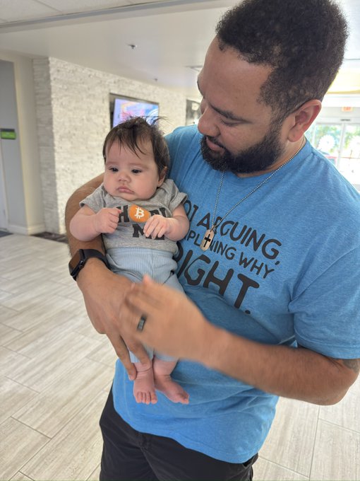 A bearded man wearing a blue t-shirt that reads 'I'M ARGUING, EXPLAINING WHY I'M RIGHT' while holding a baby wearing a Bitcoin-themed shirt.