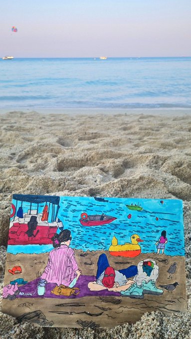 A colorful painted artwork depicting a beach scene with figures, boats, and a yellow duck rests on real sand with the ocean in the background.