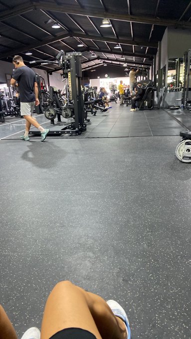 A first-person view from someone sitting on a gym floor, showing their legs in the foreground while a man in a dark shirt walks past weight racks in a spacious industrial-style gym.