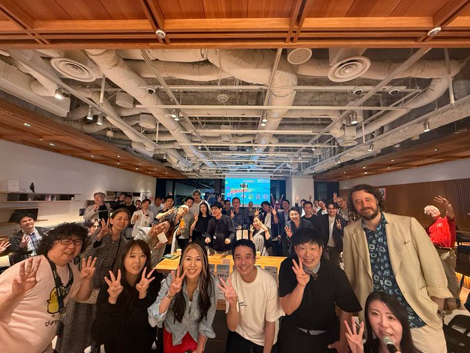 A large group of people in a modern office or event space pose for a photo, many raising peace signs toward the camera.