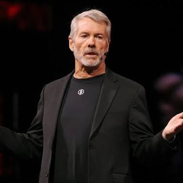 Michael Saylor, the executive chairman of Strategy (formerly MicroStrategy), stands on stage with arms outstretched, likely delivering a Bitcoin-focused presentation that aligns with the company's recent update on its paused BTC acquisitions and shift toward share buybacks.